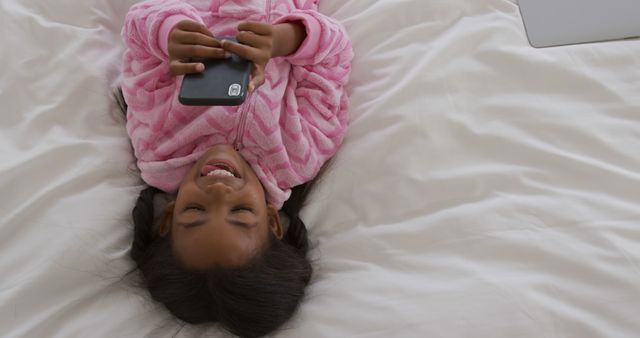 Young child relaxing in bed, smiling at mobile phone. Ideal for themes of childhood joy, technology use, and relaxation. Great for family lifestyle, parenting, and educational technology content.