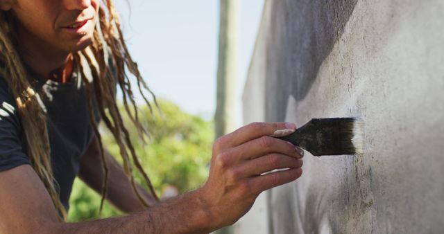 The scene shows a person with dreadlocks using a paintbrush to paint a wall outdoors under sunny weather. The image can be used in articles or advertisements about home renovation, outdoor DIY projects, artistic expression, and personal hobbies. It is suitable for websites focusing on home improvement, creativity, and lifestyle topics.