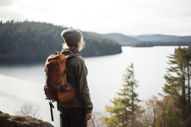Woman hiking on trail overlooking serene lake with dense forest. Suitable for topics on outdoor activities, adventure, solitude and mental well-being, or nature exploration. Perfect for travel blogs, outdoor equipment brands, adventure promotions, and wellness articles.
