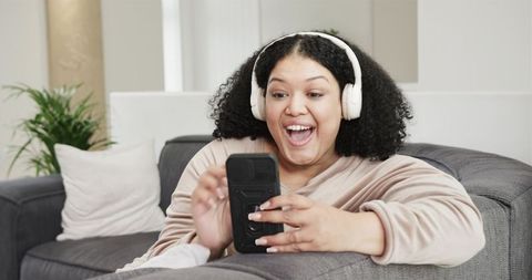 African American woman relaxing on sofa wearing headphones and using smartphone at home