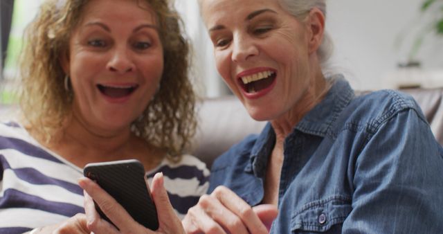Senior women smiling and using a smartphone together at home. Perfect for themes about friendship, technology use among elderly, leisure activities, and happy moments in daily life.