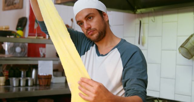 A chef wearing a white hat and blue shirt is in a professional kitchen stretching dough. Use this image for culinary content, restaurant promotions, cooking blog or social media posts focusing on food preparation, professional cooking techniques, or culinary arts.