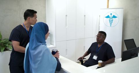 Diverse clinic team talking at reception holding coffee wearing navy scrubs and badges