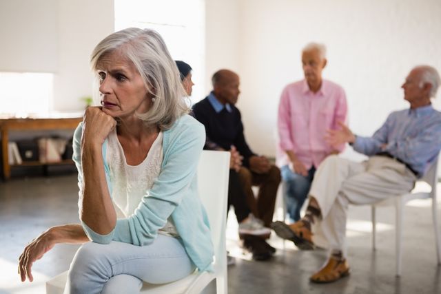 Senior woman looking worried while sitting in a group setting with friends in the background. Ideal for use in articles or advertisements about senior care, mental health, community activities, or aging.