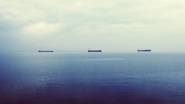 Three cargo ships are floating on a calm sea under a cloudy sky. This serene and tranquil scene highlights the vastness of the ocean and the peacefulness of maritime transport. This scene can be used in contexts related to shipping, transport industry, maritime trade, and nature photography. Suitable for illustrating concepts of solitude, vastness, and global trade.