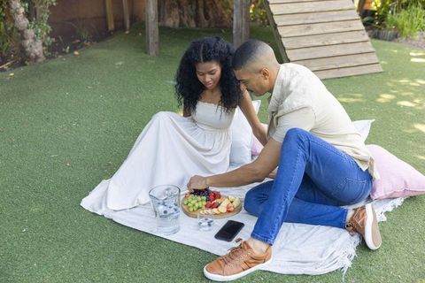Couple sharing fruit platter on picnic blanket in sunny backyard garden, relaxing