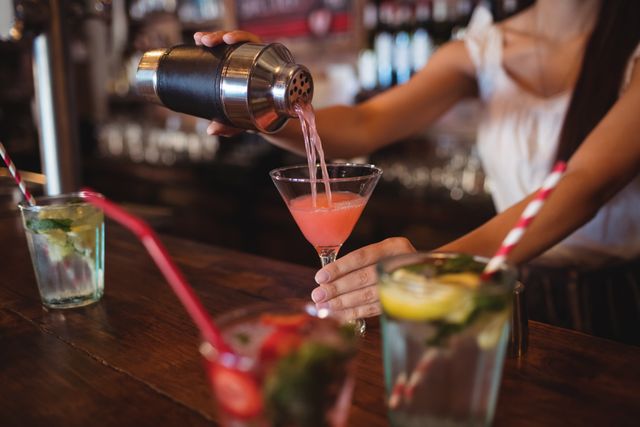 Mid section view of female bartender pouring pink cocktail into a glass on bar counter. Other colorful drinks with straws are present, suggesting a busy and festive bar environment. Ideal for use in promotions related to nightlife, bar services, cocktail recipes, or event advertisements.