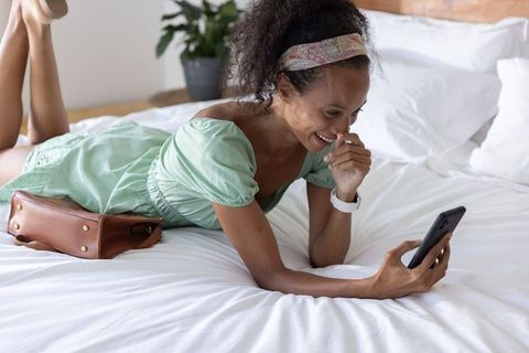 Smiling woman lying on bed using smartphone for messages, casual home relaxation
