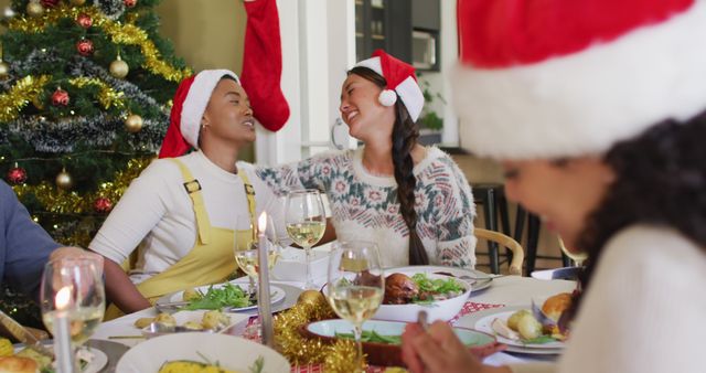 Diverse group of friends celebrating Christmas at a festive dinner table. Wearing Santa hats, they are sharing a joyous moment with laughter and conversation. The setting includes a beautifully decorated Christmas tree, festive ornaments, and a delicious meal with drinks. Ideal for holiday greeting cards, festive advertisements, and promotional materials focusing on togetherness and holiday spirit.