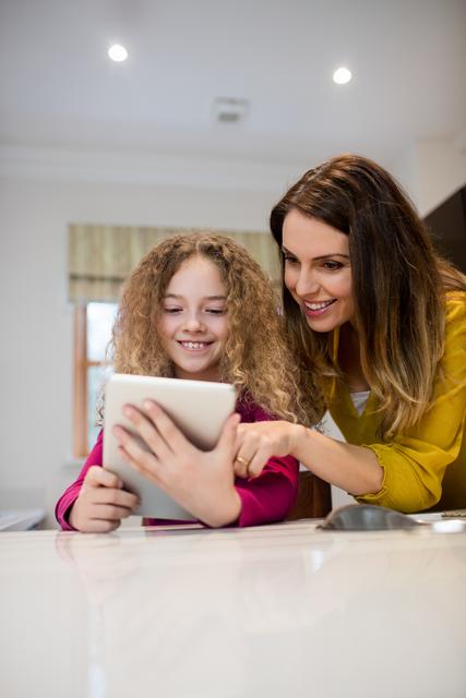 Mother and daughter are smiling while using a digital tablet together in the kitchen. This image can be used for family-oriented advertisements, parenting blogs, technology in family life, and educational content.