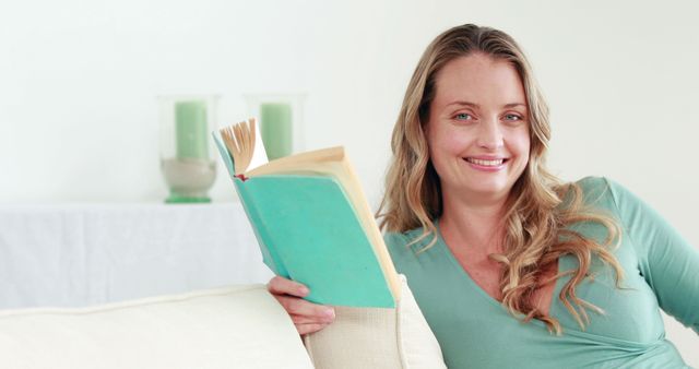 Smiling woman reading book while comfortably sitting on sofa at home. Calm and relaxed atmosphere with green accents in background. Ideal for illustrating concepts of leisure, relaxation, home lifestyle, education, and personal time.