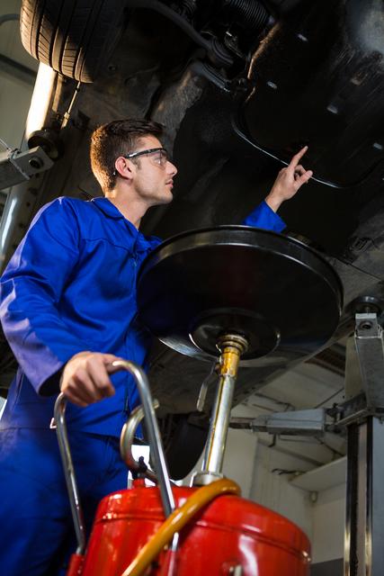 Mechanic in blue uniform servicing a car in an auto repair garage. He is inspecting the vehicle's undercarriage and using tools under a lifted car. This image can be used for themes related to car maintenance, auto repair services, mechanical work, professional technicians, and automotive industry advertisements or tutorials.