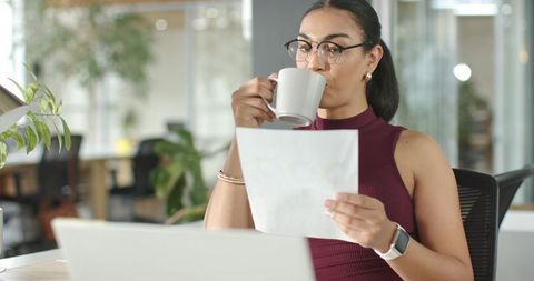 Professional woman sipping coffee while reading document at open-plan office desk