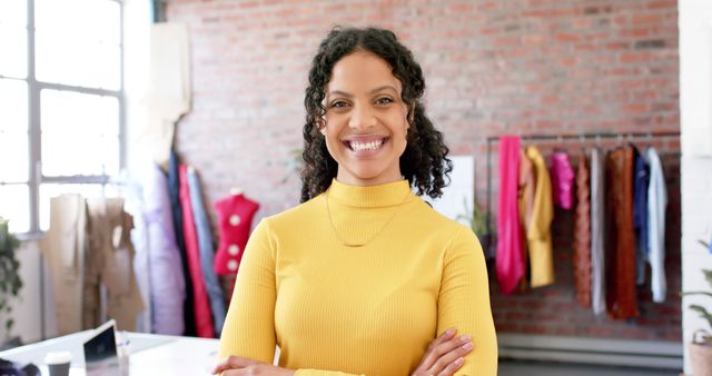 Fashion designer smiling confidently while standing in a modern studio with textiles and clothing in the background. Ideal for content related to fashion design, creative professions, entrepreneurship, diversity in business, and modern workspaces.