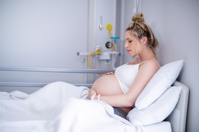 Pregnant woman in hospital gown lying on a bed in a hospital room. She appears calm and relaxed, suggesting a moment of rest before childbirth. This image can be used for topics related to maternity care, hospitals, prenatal health, and expecting mothers. Ideal for use in healthcare brochures, maternity ward advertisements, and pregnancy articles.