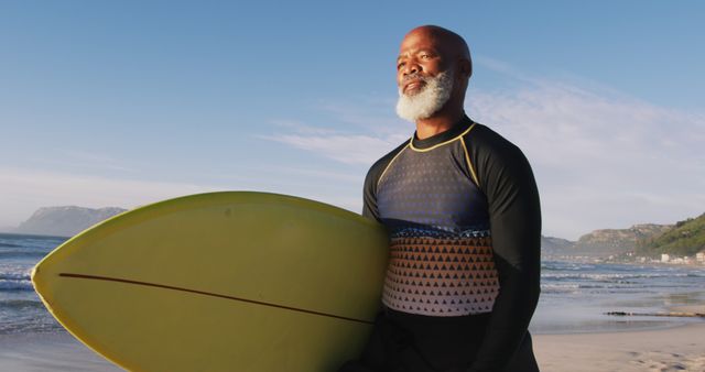 Senior man holding surfboard while standing on beach during sunset. He is dressed in a wetsuit, looking at the ocean, embodying active retirement and healthy lifestyle. Ideal for depicting themes of fitness, healthy aging, adventure, and outdoor activities for seniors.