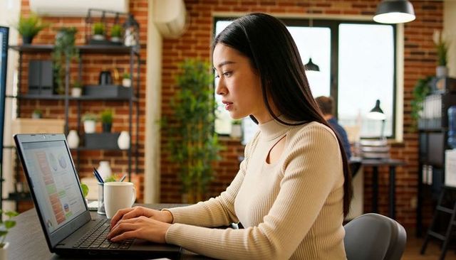 Asian woman typing and reviewing laptop charts in modern exposed brick coworking space