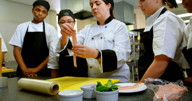 Group of student chefs attentively watching their instructor demonstrate preparation techniques in a professional kitchen. This detailed shot can be used for educational materials, cooking school promotions, culinary arts programs advertisements, and food industry articles, showcasing hands-on learning and skill development in the culinary field.