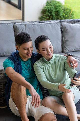 Couple relaxing in activewear sharing smartphone outdoors on rattan sofa near garden
