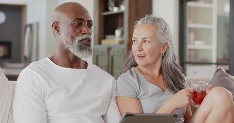 Diverse senior couple relaxing on sofa with tablet and tea in modern open-plan living room