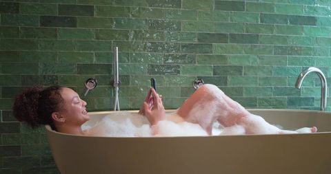 African american woman relaxing in bubble bath using smartphone in green-tiled bathroom