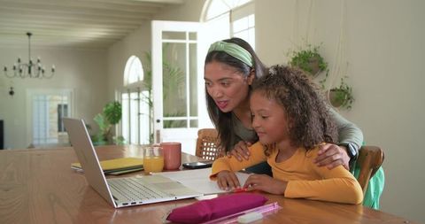 Mother guiding daughter doing homework at wooden table using laptop and worksheets