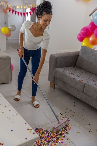 Smiling teen girl sweeping metallic confetti after birthday party in bright living room