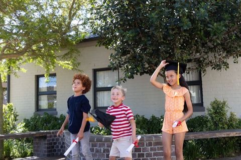 Diverse children celebrating kindergarten graduation outdoors holding caps and diplomas