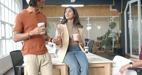 Diverse coworkers chatting in modern open-plan office over coffee, collaborating on laptop