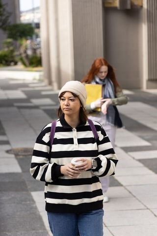 Hispanic student walking on urban campus holding coffee and yellow folder wearing beanie