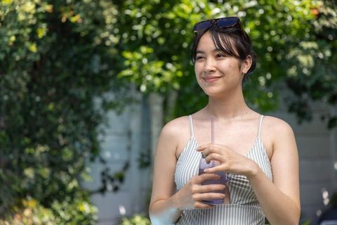 Sunlit asian woman smiling in striped sundress holding purple drink outdoors wearing sunglasses