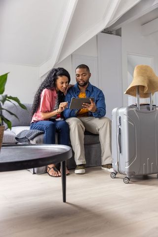Black couple planning trip on tablet in modern loft with luggage and straw hat