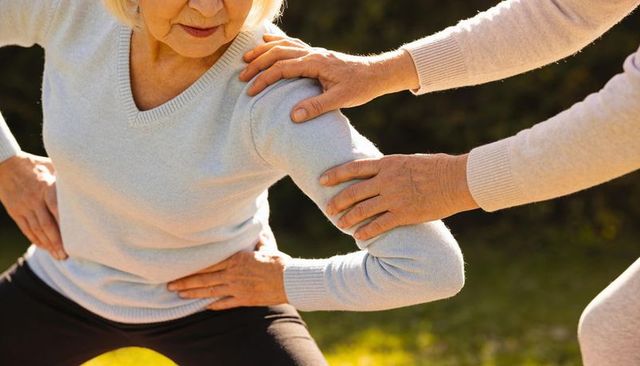 Senior woman performing assisted squat outdoors with caregiver hands supporting balance