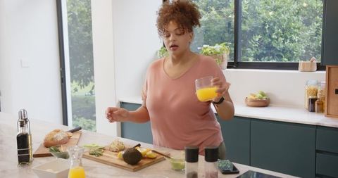 Woman Preparing Avocado Toast While Holding Orange Juice in Bright Modern Kitchen