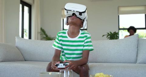 African American boy playing virtual reality game wearing VR headset and controller on living room c