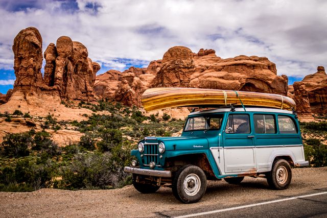Vintage SUV with a canoe on top is parked on a roadside with majestic red rock formations in the background under a partly cloudy sky. Perfect for conceptualizing adventure travel, road trips, outdoor activities, exploration themes. Ideal for use in travel agencies, automotive advertisements, adventure blogs, and landscape posters.