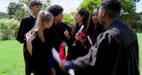 Group of graduates celebrating on campus lawn holding diplomas with red ribbons
