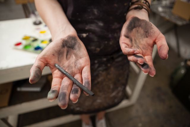 Close-up of female artist's hands covered in charcoal, holding a charcoal pencil. Ideal for use in articles or advertisements about art, creativity, and the artistic process. Suitable for illustrating the hands-on nature of artistic work and the tools used by artists.