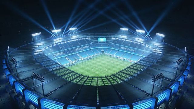 Illuminated oval stadium under night lights, floodlights highlighting empty green pitch