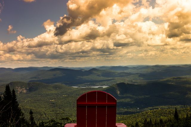 A lone red chair sits at the edge, offering a panoramic view of a mountainous landscape under a sky filled with scattered clouds. This serene and peaceful setting evokes thoughts of reflection and solitude. Ideal for travel magazines, nature conservation campaigns, motivational posters, or any project highlighting the beauty and tranquility of untouched nature.