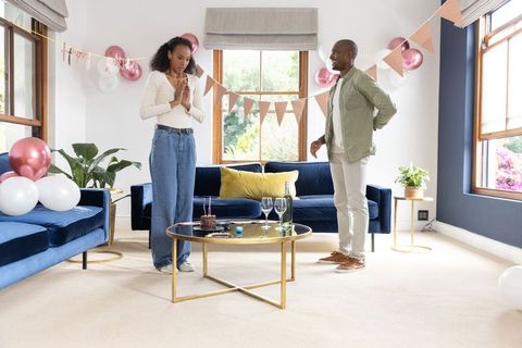 Couple celebrating birthday surprise in bright living room with cake, balloons, wine