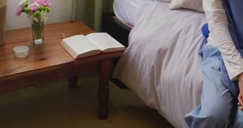 Woman sitting on bed reading beside wooden nightstand with vase of flowers cozy bedroom scene