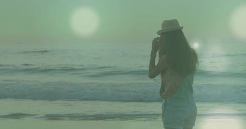 Woman standing on beach holding straw hat gazing at ocean sunset with soft lens flare