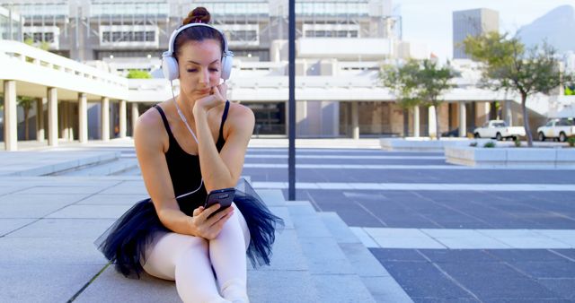 Caucasian woman sits outdoors in a cityscape, with copy space. She's a ballet dancer taking a break, enjoying music on her headphones.