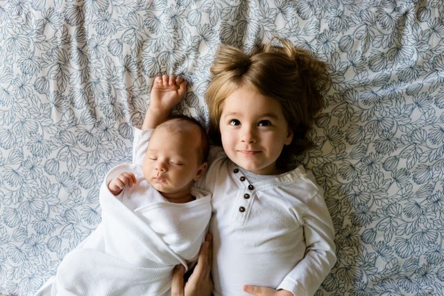 Two young siblings, a newborn baby and an older toddler, lying in bed together and looking up. They are wearing white clothes, and the baby is wrapped in a swaddle. This image is ideal for use in parenting blogs, family-oriented advertisements, and baby product promotions. The overhead perspective provides a warm feel and highlights the innocence and bond between the siblings.