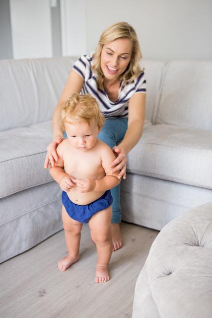 Mother helping her baby boy take his first steps in a cozy living room. Ideal for use in parenting blogs, family-oriented advertisements, child development articles, and home lifestyle promotions.