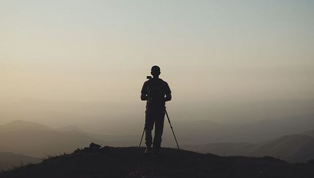 Silhouette photographer standing on mountain ridge framing camera on tripod at sunrise