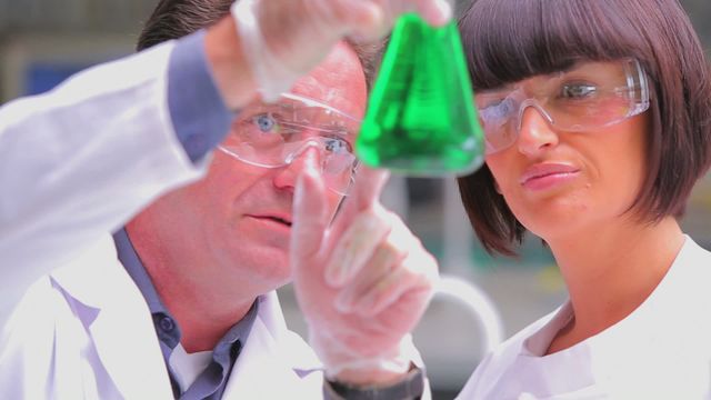 Two chemists are analyzing a green liquid in a laboratory setting. Dressed in white lab coats, safety goggles, and gloves, they carefully observe the contents of the beaker, emphasizing the collaborative nature of scientific research. Perfect for illustrating teamwork in science, educational materials, or scientific discoveries.