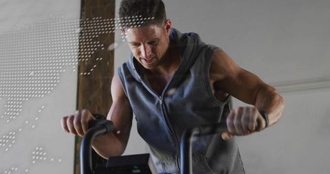 Fit man performing intense stationary bike workout in sleeveless hoodie, wristband visible