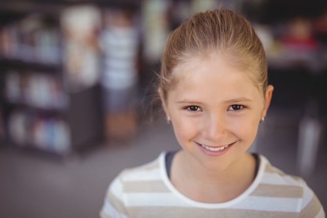 This image shows a young schoolgirl smiling in a library, making it ideal for educational content, school promotions, and child development articles. It can be used in advertisements for educational programs, library services, or children's books. The cheerful expression and academic setting convey a positive learning environment.